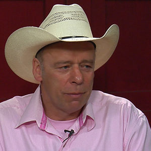 Carl Cosack, in a pink shirt and tan hat, doing an interview with a red wall in the background.