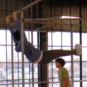 Someone swinging on an overhead bar, while another person walks by in the background.